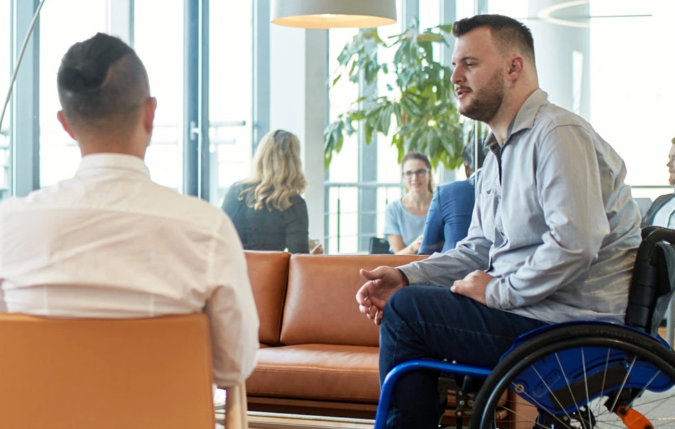 Foto de uma sala com parede de janelas brancas, sofá e poltrona terracota, lustre branco em semi-lua com luz amarela e plantas em vasos no chão. Um homem de blusa branca está sentado na poltrona de costas para a câmera e ao lado dele um homem de cadeira de rodas conversa com ele, vestido de blusa social cinza e calça jeans azul. 